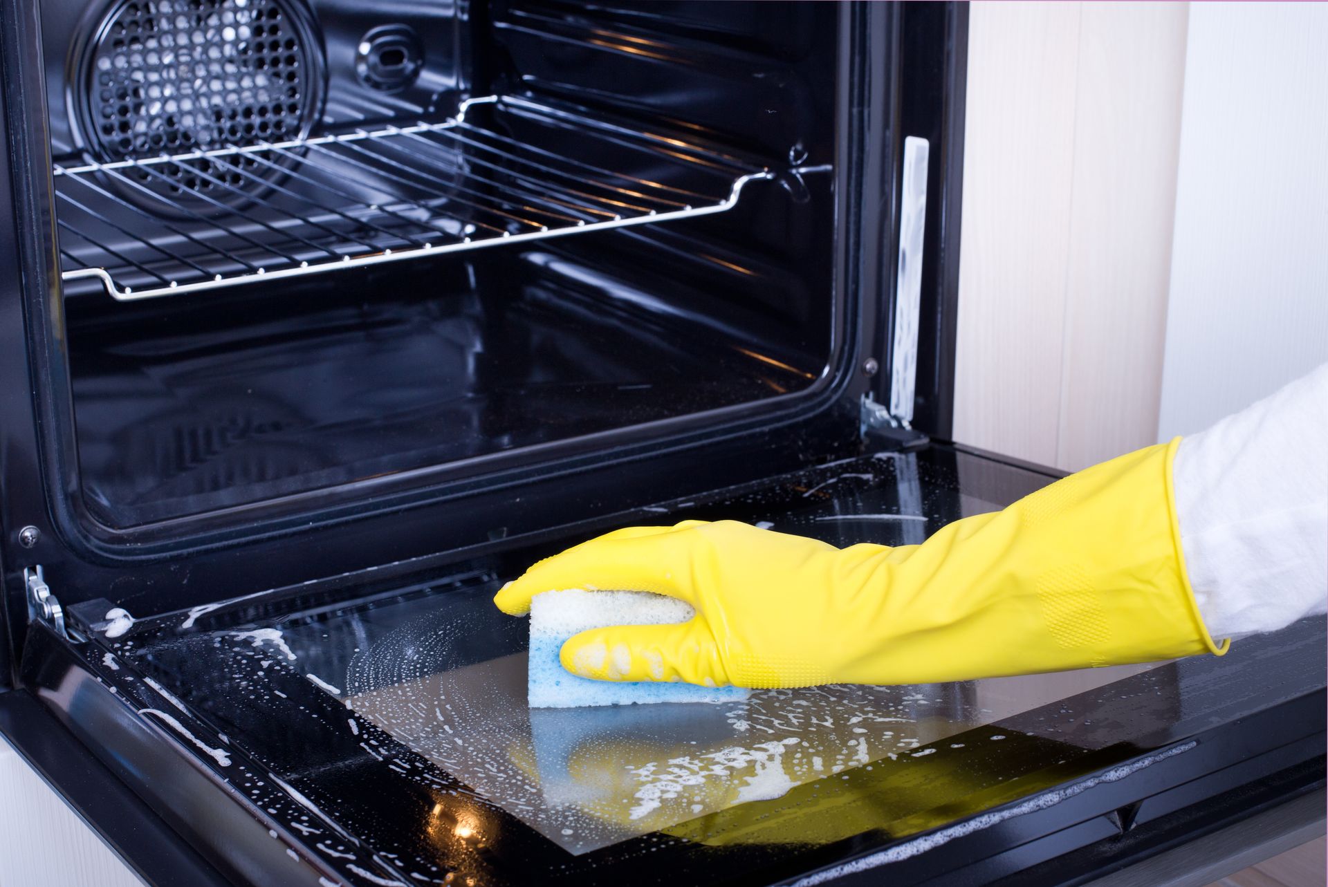 A person wearing yellow gloves is cleaning an oven with a sponge.