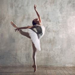 A ballerina is dancing on one leg in front of a wall.
