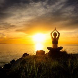 A woman is sitting in a lotus position in front of the ocean at sunset.