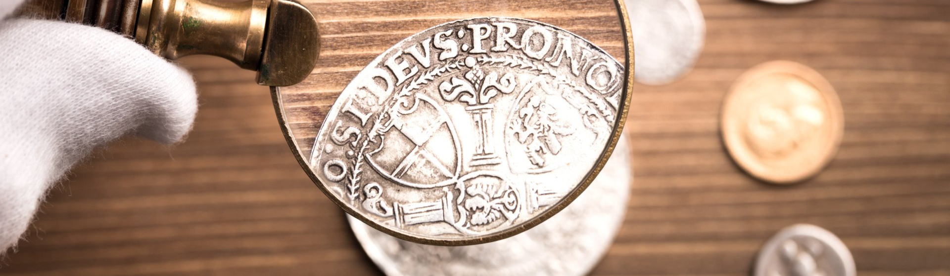 Hand holding a magnifying glass examining a coin on a table with 5 other coins