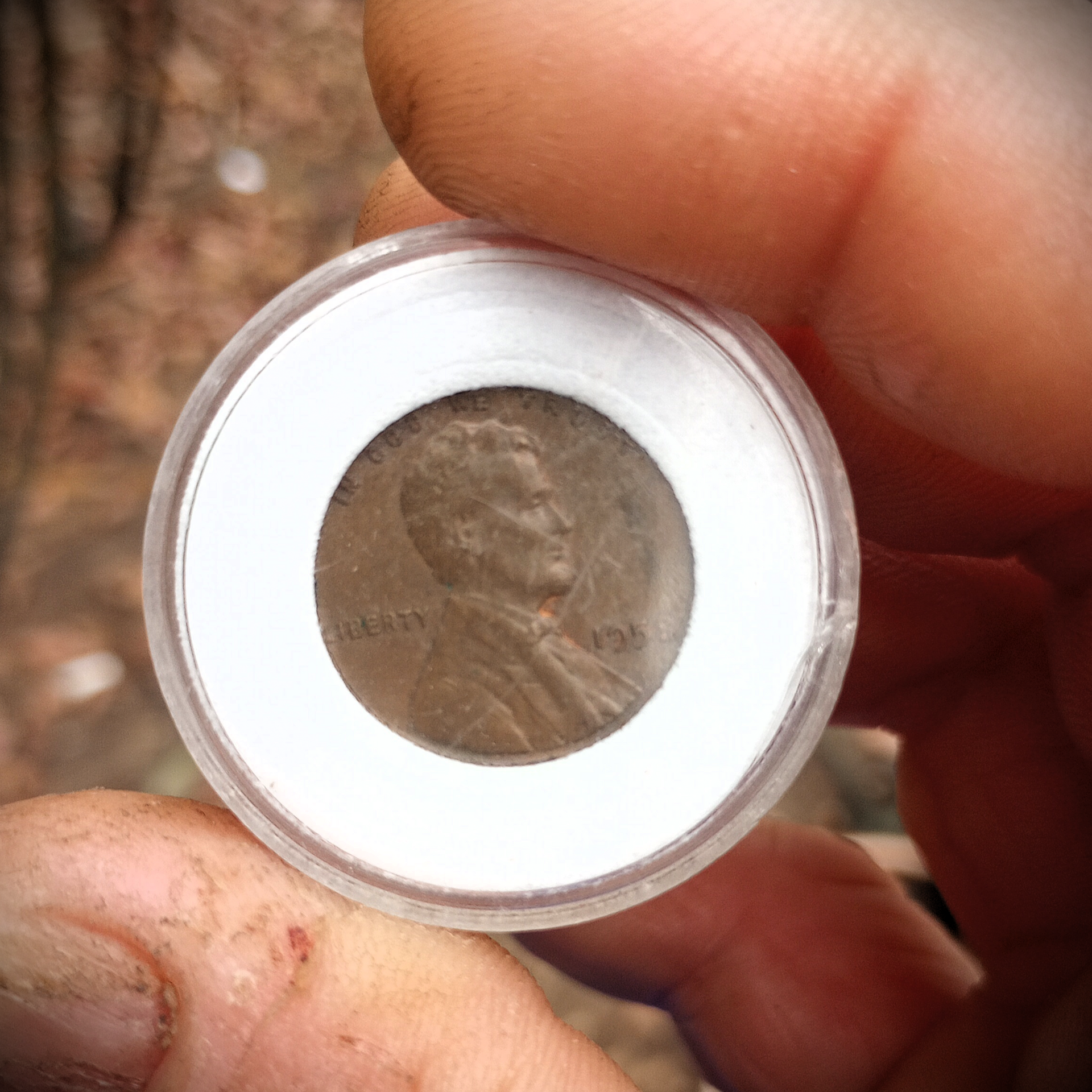 A coin in a clear plastic holder, held between fingers, with a worn profile visible in the center.