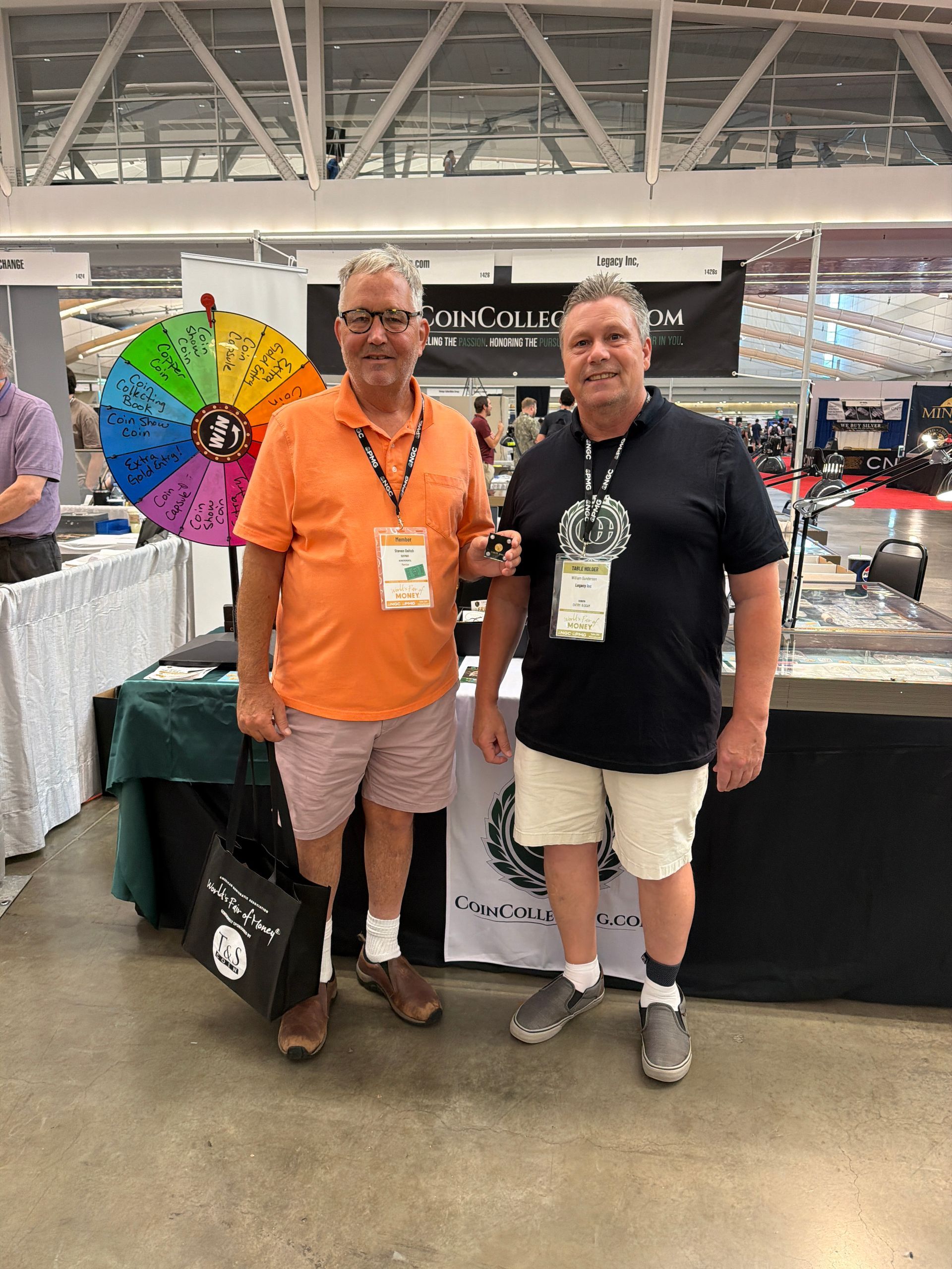Two men posing at a convention booth beside a colorful prize wheel