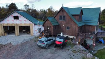 Rustic house and garage with teal roofs, parked cars, and a red utility vehicle on a gravel driveway.