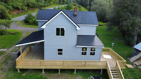 Gray house with blue roof and wraparound porch on a wooded hillside, viewed from above
