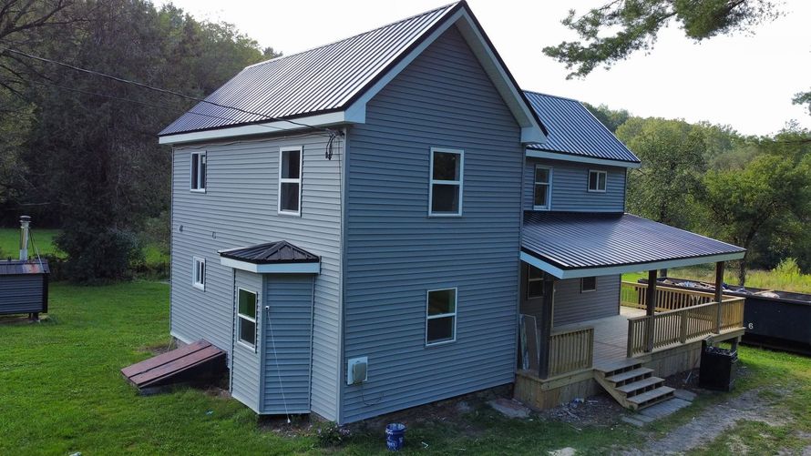 Blue two-story house with a metal roof and covered porch in a grassy wooded yard