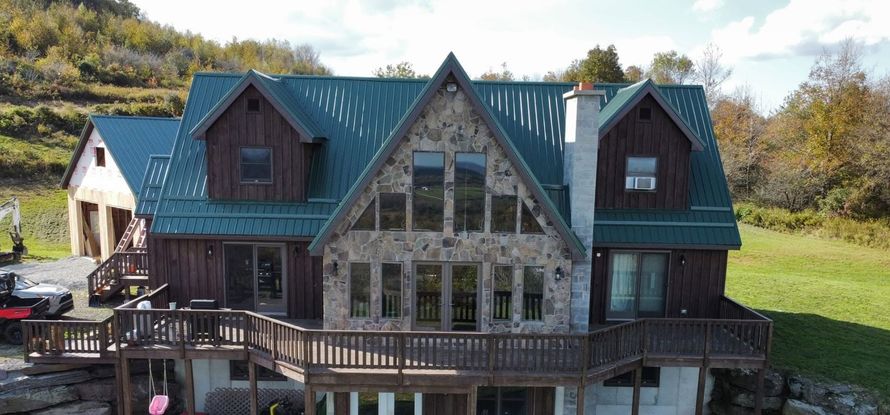 Rustic house with green metal roof, dormer windows, and wooden deck in a sunny hillside setting
