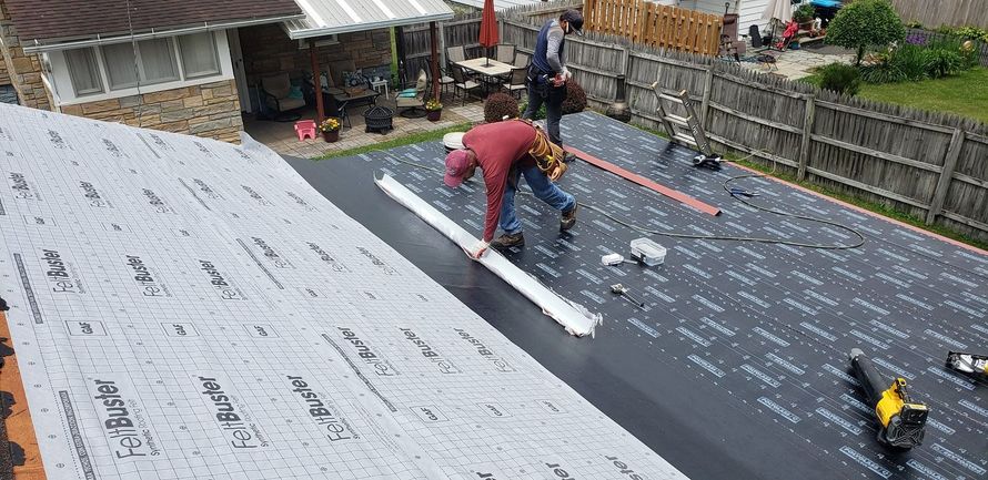 Two workers installing black roofing on a sloped roof beside a house and fence