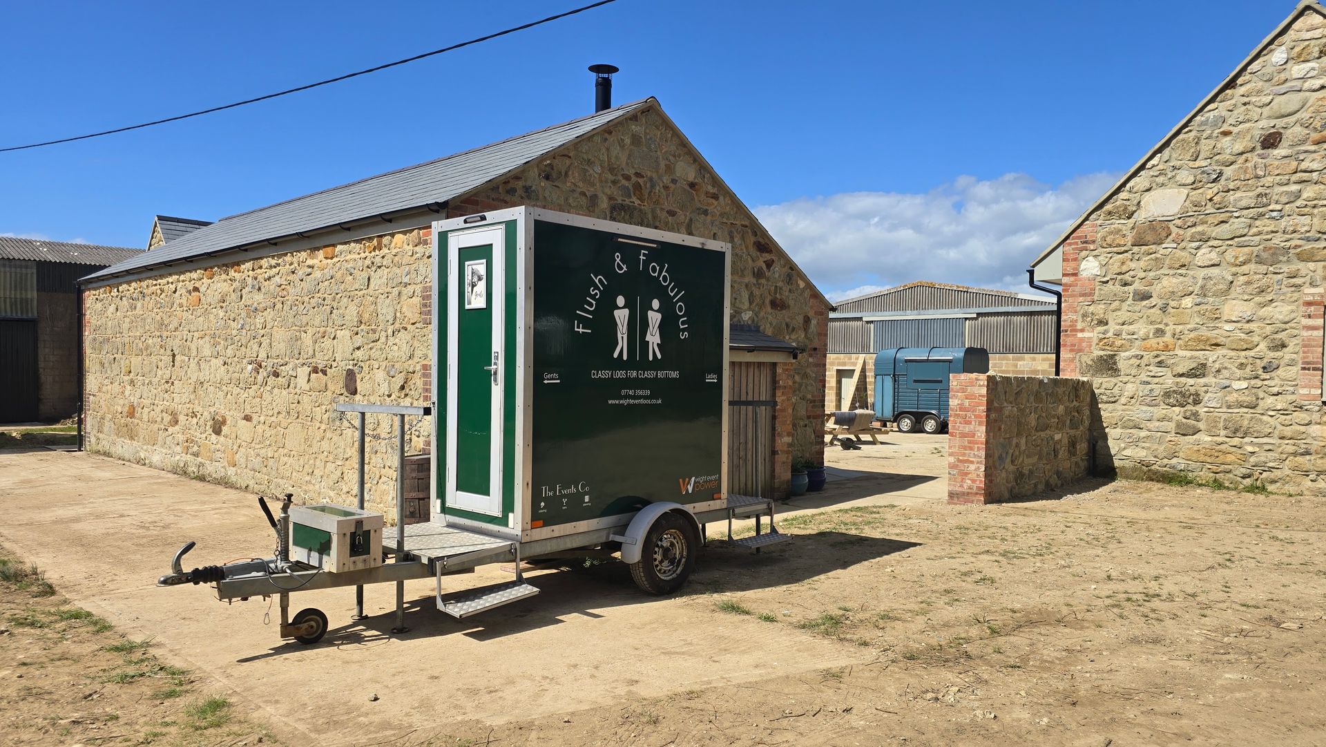 Portable toilet trailer beside stone buildings in a sunny rural yard