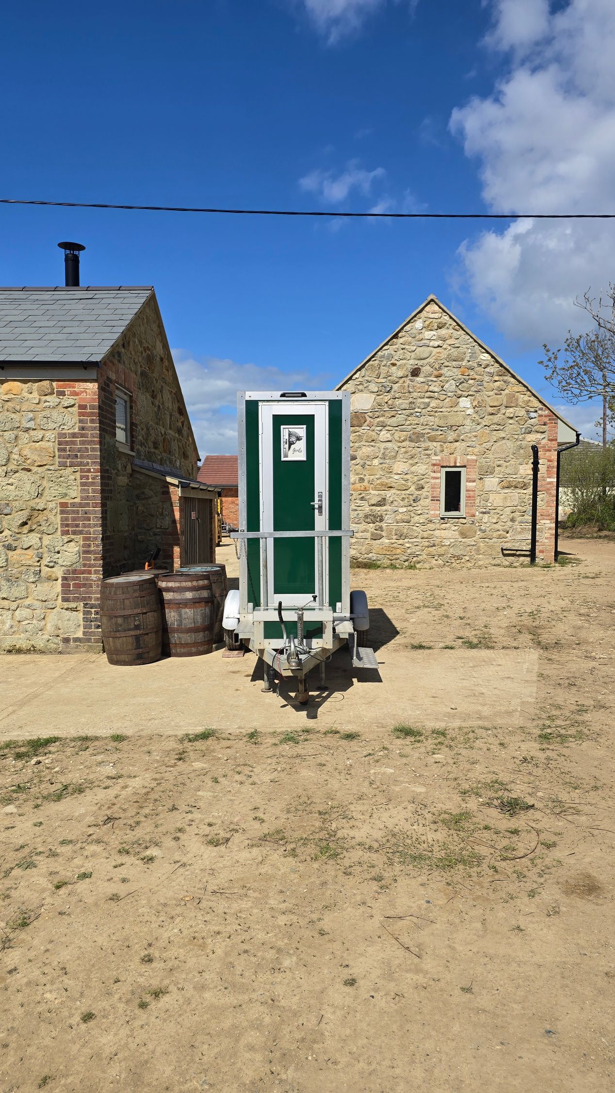 Two small stone buildings with a green door, barrels, and a bench on a sunny sandy yard under blue sky
