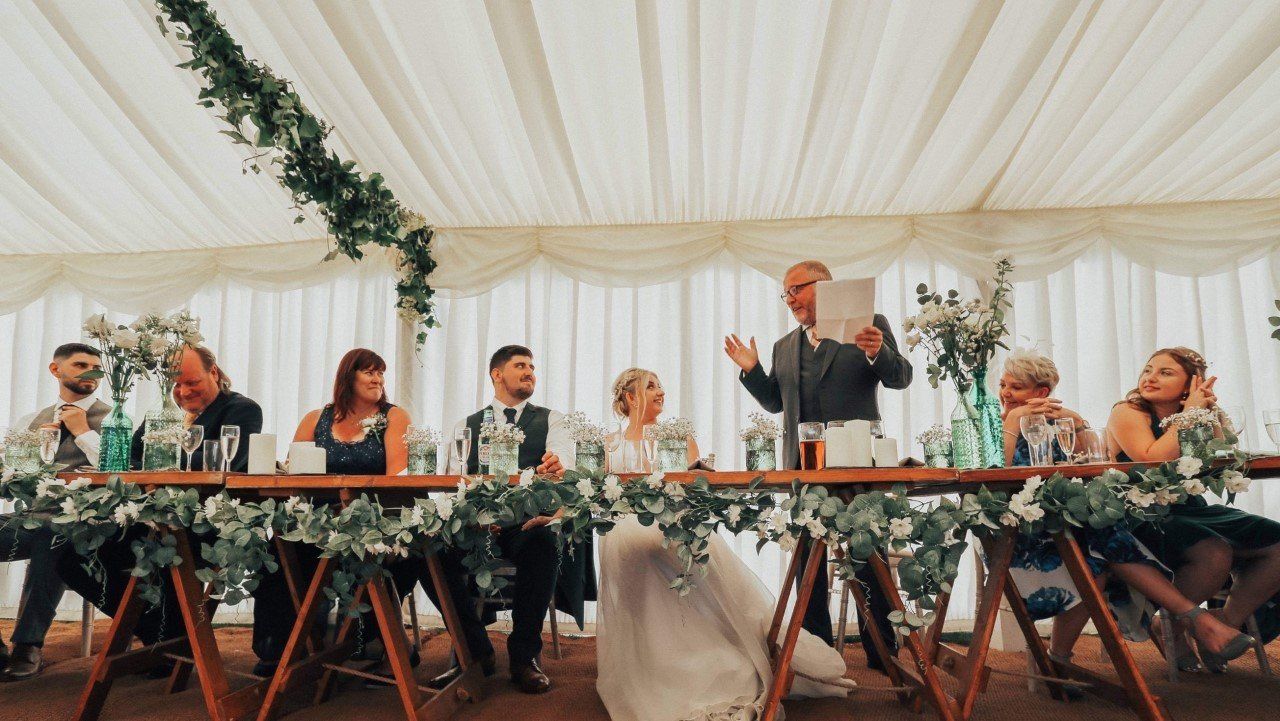 A man is giving a speech at a wedding reception in a tent.
