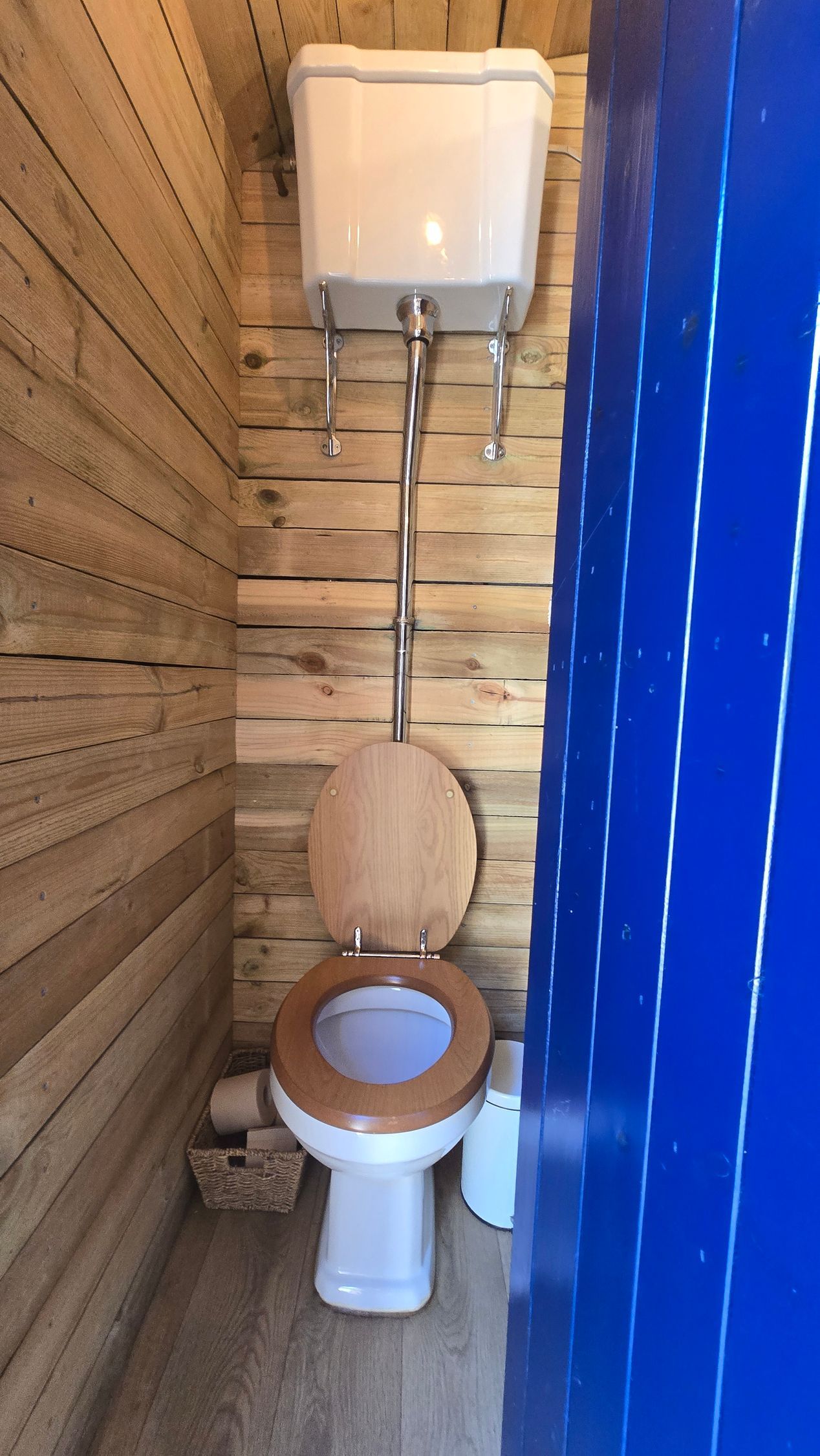 Toilet with wooden seat in a narrow rustic bathroom beside a blue shower curtain