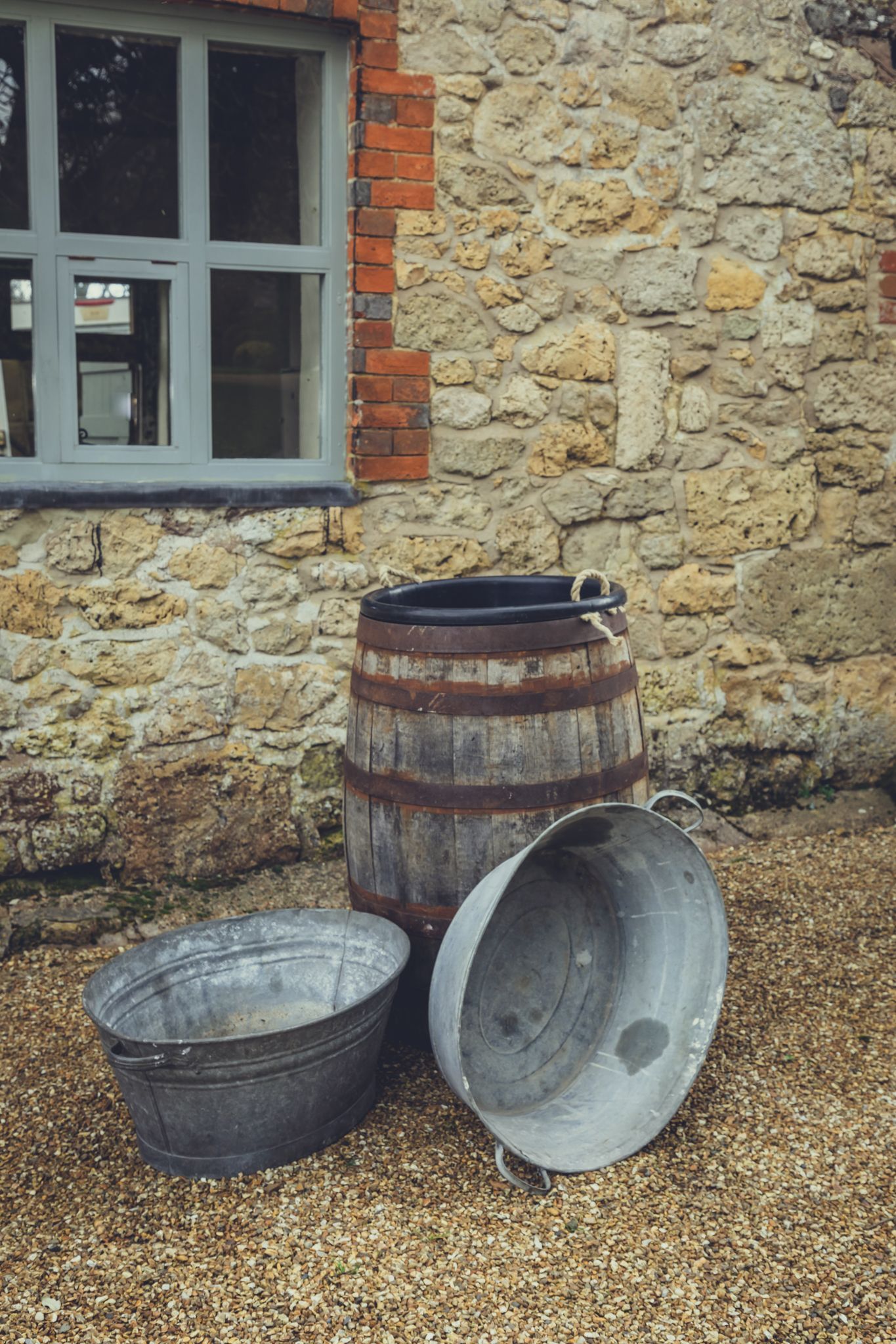 Three metal buckets and a wooden barrel are sitting on gravel in front of a stone building.