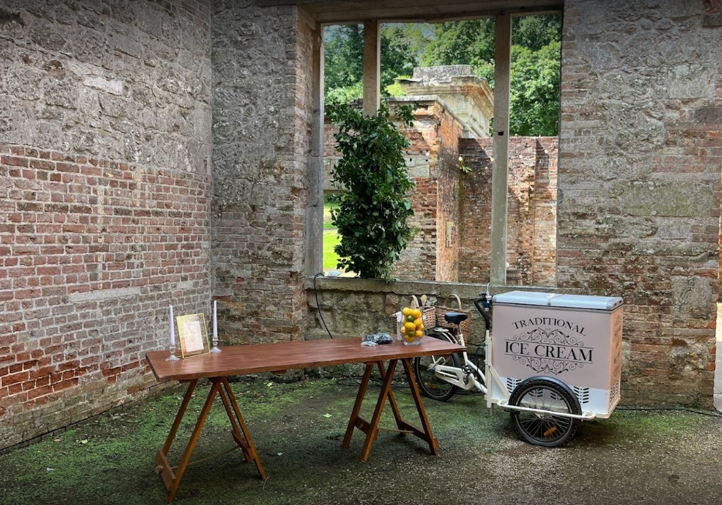 An ice cream cart is parked next to a wooden table in front of a brick wall.