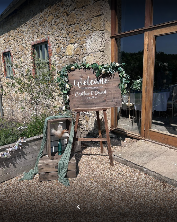 A wooden welcome sign is sitting on a wooden easel in front of a stone building.
