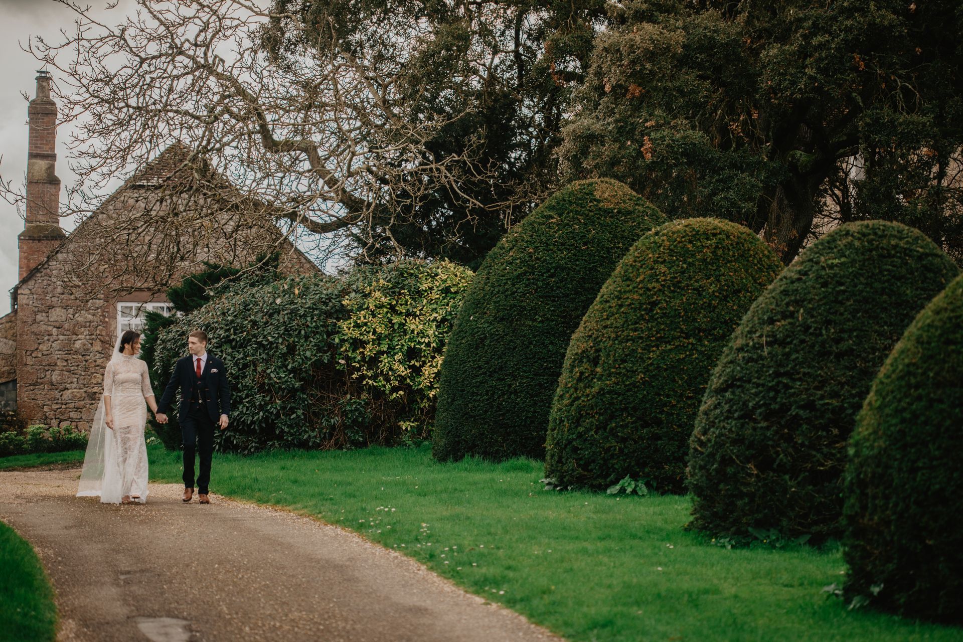 A bride and groom are walking down a path in front of a stone building.