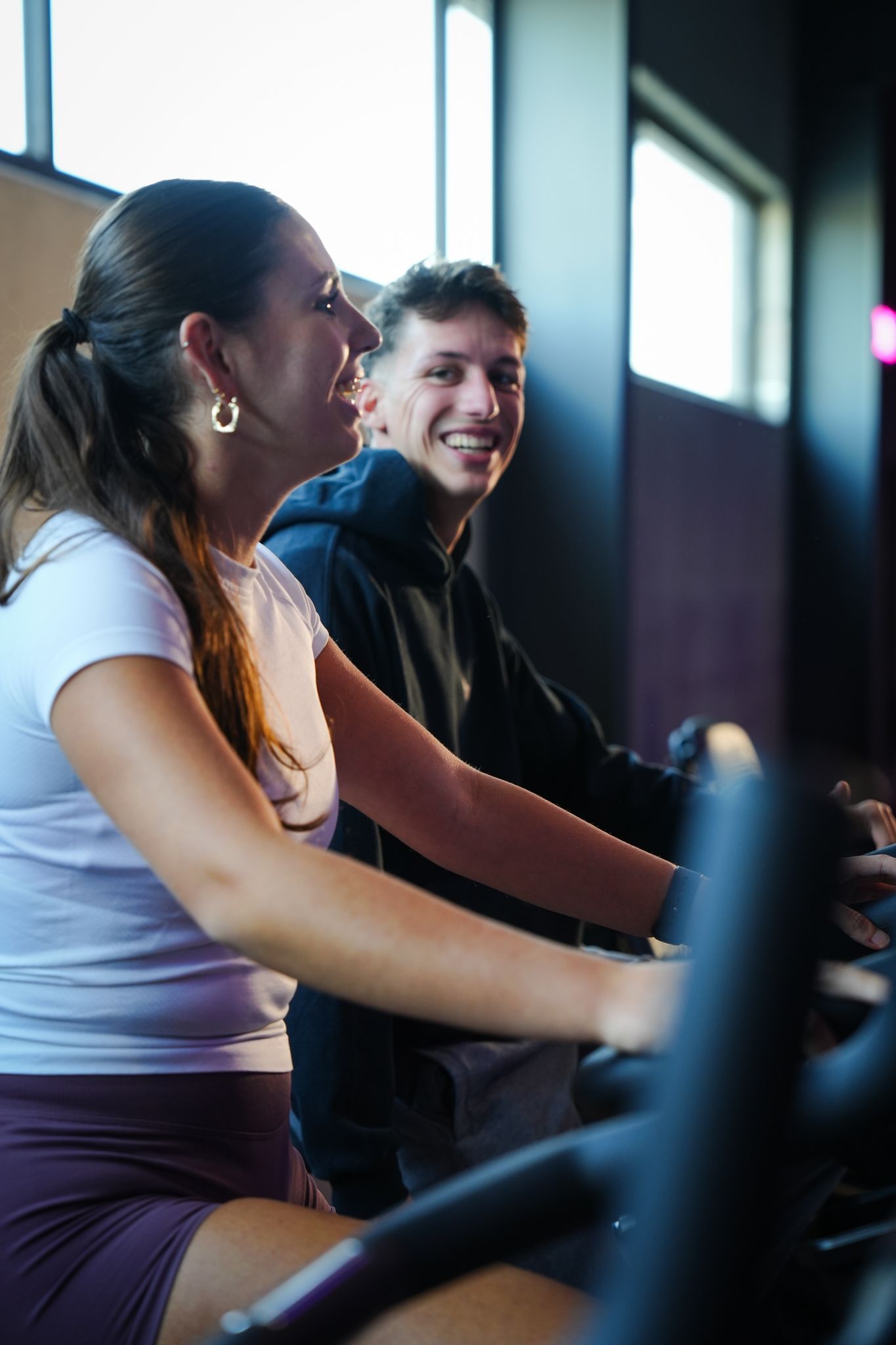 Een vrouw en een man lachen terwijl ze op een hometrainer fietsen in een sportschool. Ze zijn goed verlicht en hebben een paars-zwarte kleurstelling.