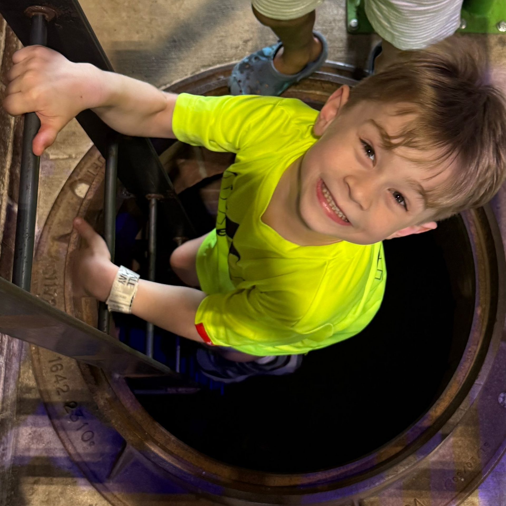 Person in a neon yellow shirt climbing a metal ladder into a dark circular opening, smiling up at the camera.