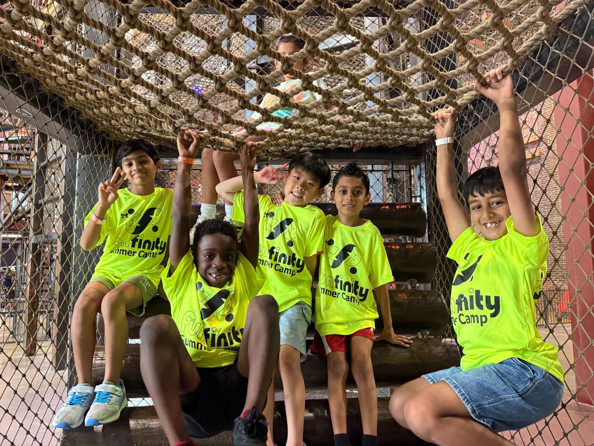 Children in neon yellow shirts smiling and hanging from a rope net in a playful indoor play area