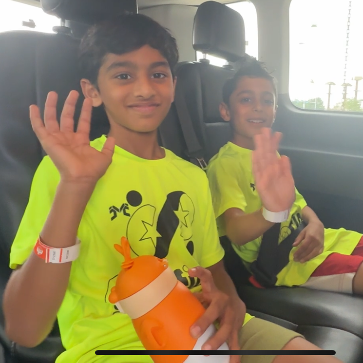 Two kids in neon shirts waving from a car seat, one holding an orange water bottle.