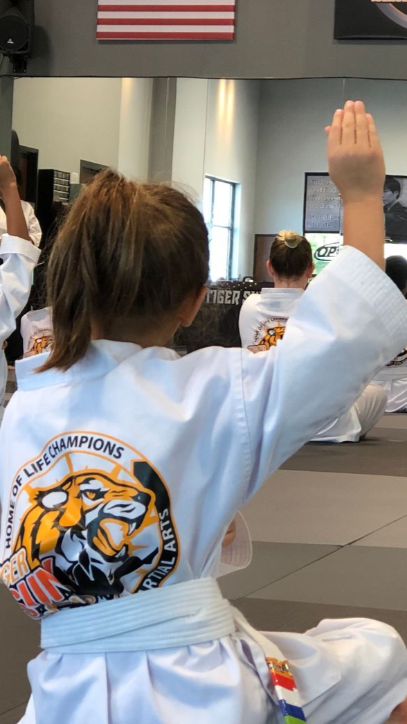 Child in a white martial arts uniform raises one arm in a dojo, with classmates and a U.S. flag in the background