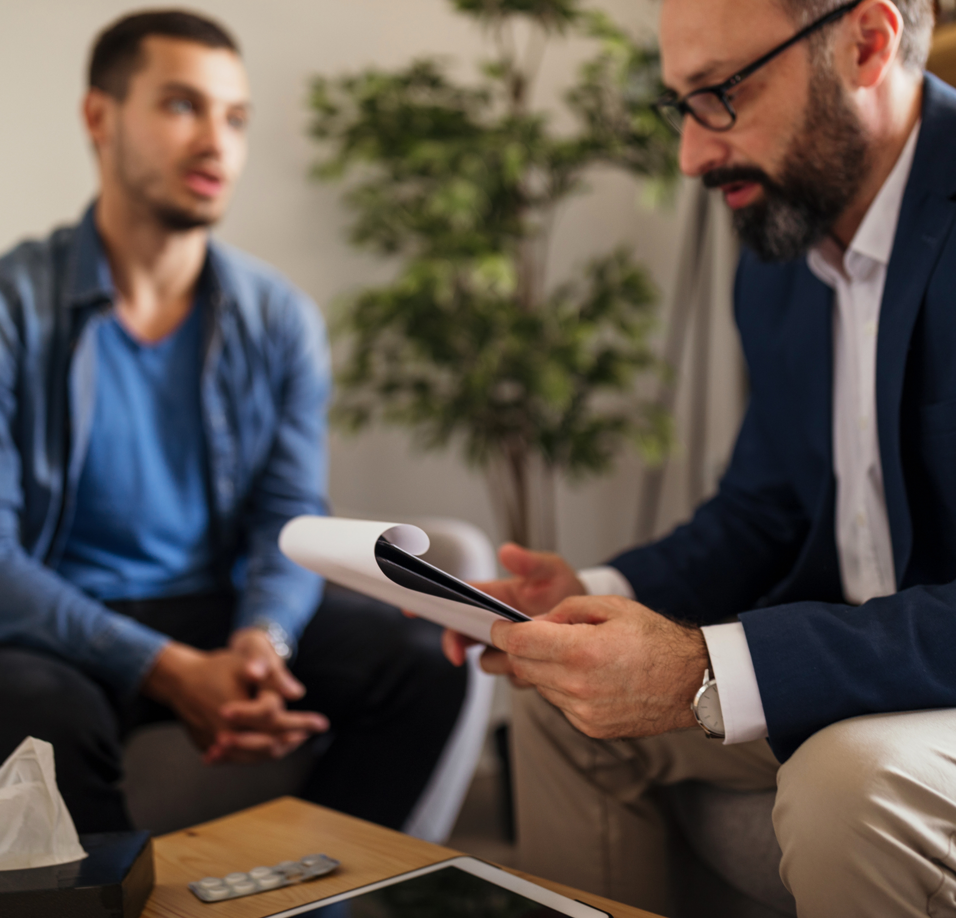 A professional in a suit reviews notes while consulting with a person in a blue shirt in a softly lit office.