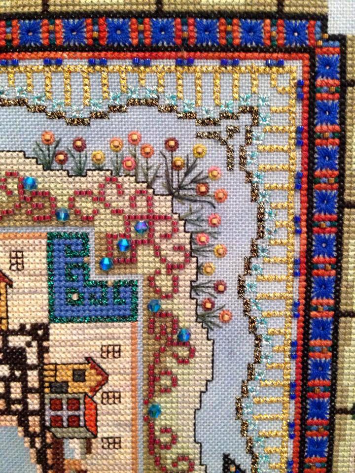 A close-up of a complex cross-stitch project featuring a floral border, brick motifs, and colorful beads on blue fabric.