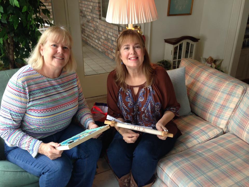 Two people sit together on a sofa holding embroidery hoops while smiling in a living room.