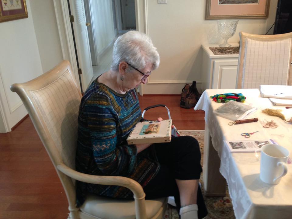 An individual with white hair sits at a dining table, working on a small embroidery project in a bright room.