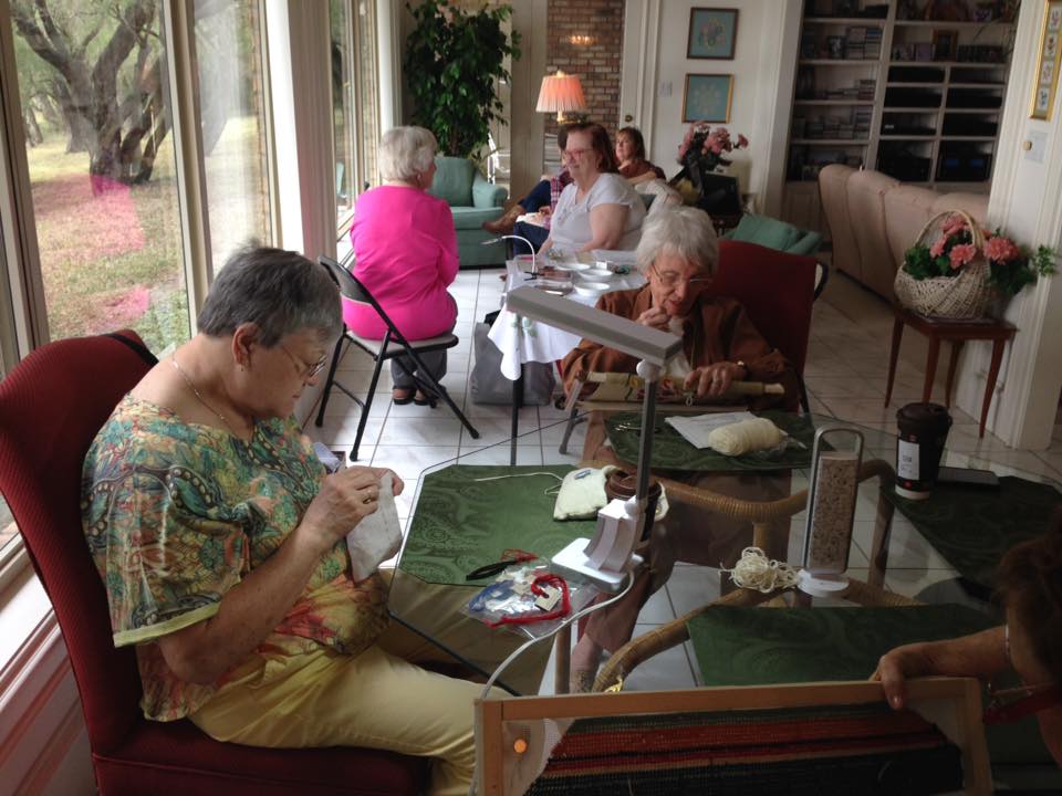Several people sit around tables in a sunlit room, focusing on embroidery work with frames and magnifying lamps.