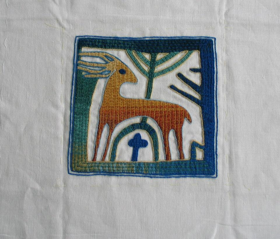 A square textile embroidery featuring a stylized, gold-toned deer, a blue cross, and a green tree on a white background.