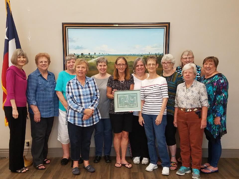 A group of thirteen people standing in a row in front of a landscape painting indoors, one person holding a framed award.