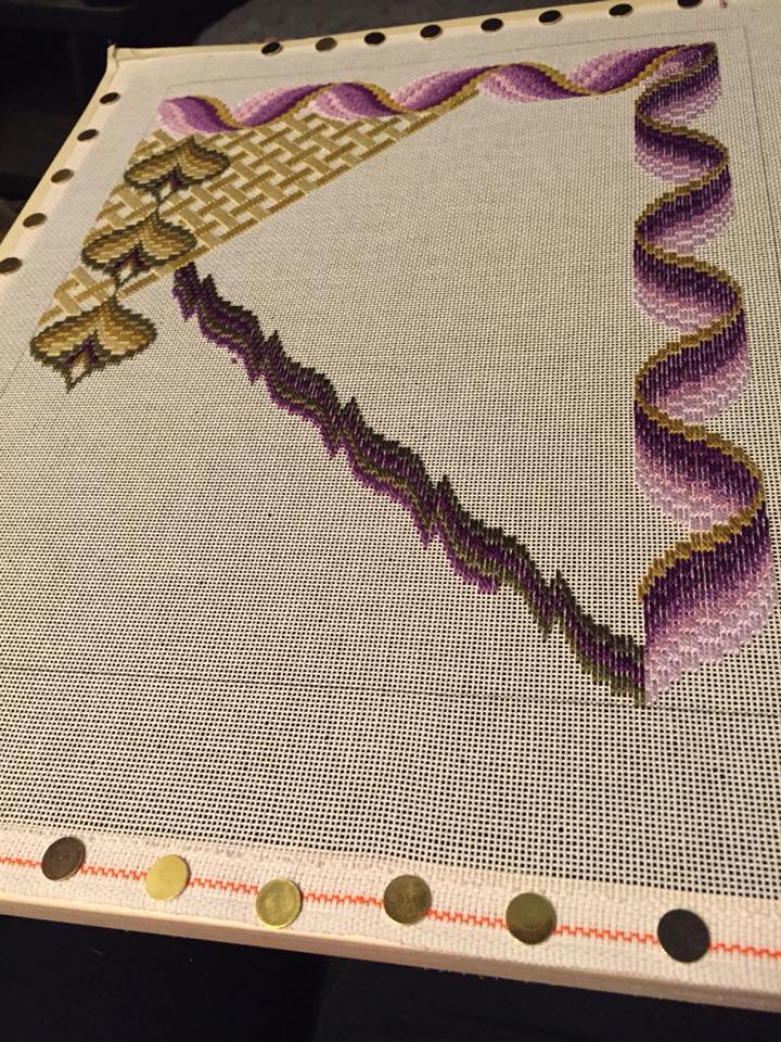 An embroidery project in progress on a frame, featuring a decorative purple ribbon border and a gold woven pattern corner.