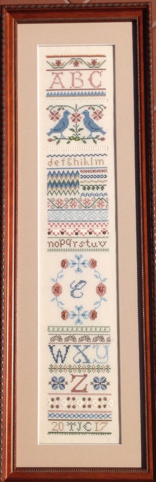 A vertical sampler featuring alphabet letters, blue birds, floral motifs, and decorative borders in blue, red, and tan.