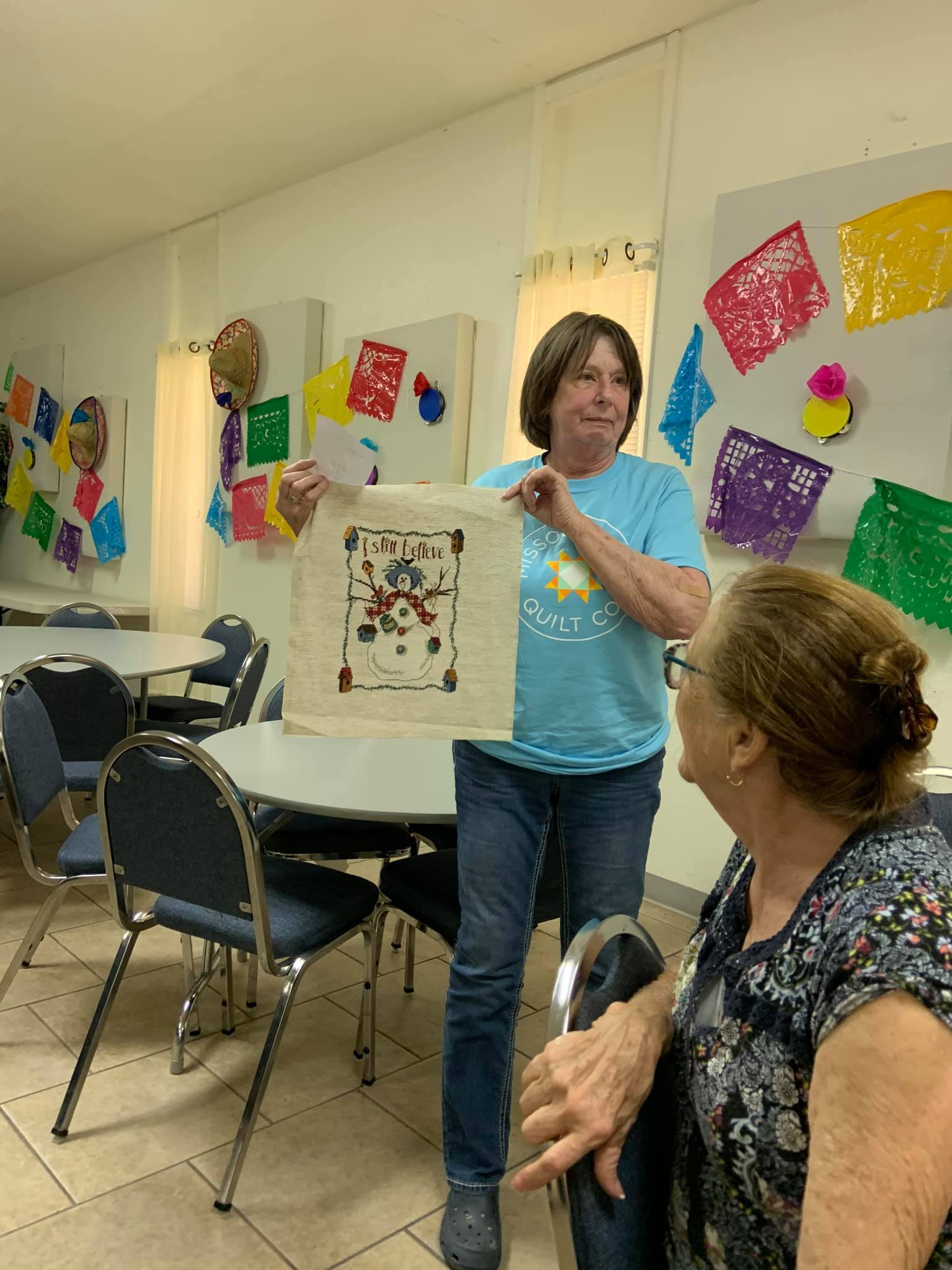 A person in a blue shirt holds up a piece of embroidered fabric for a seated person in a room with wall decorations.