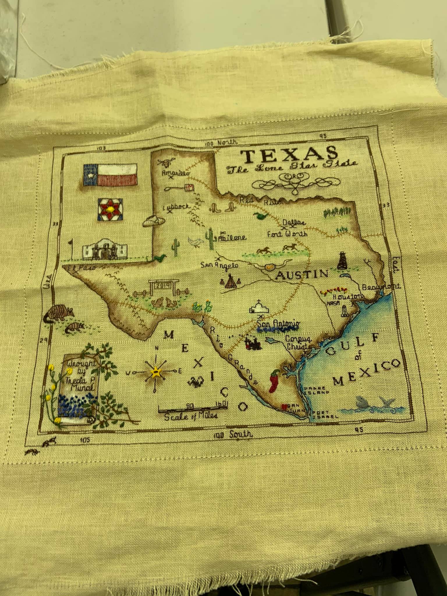 A cross-stitch embroidery map of Texas on light fabric, featuring the state flag, symbols, and labels for major cities.