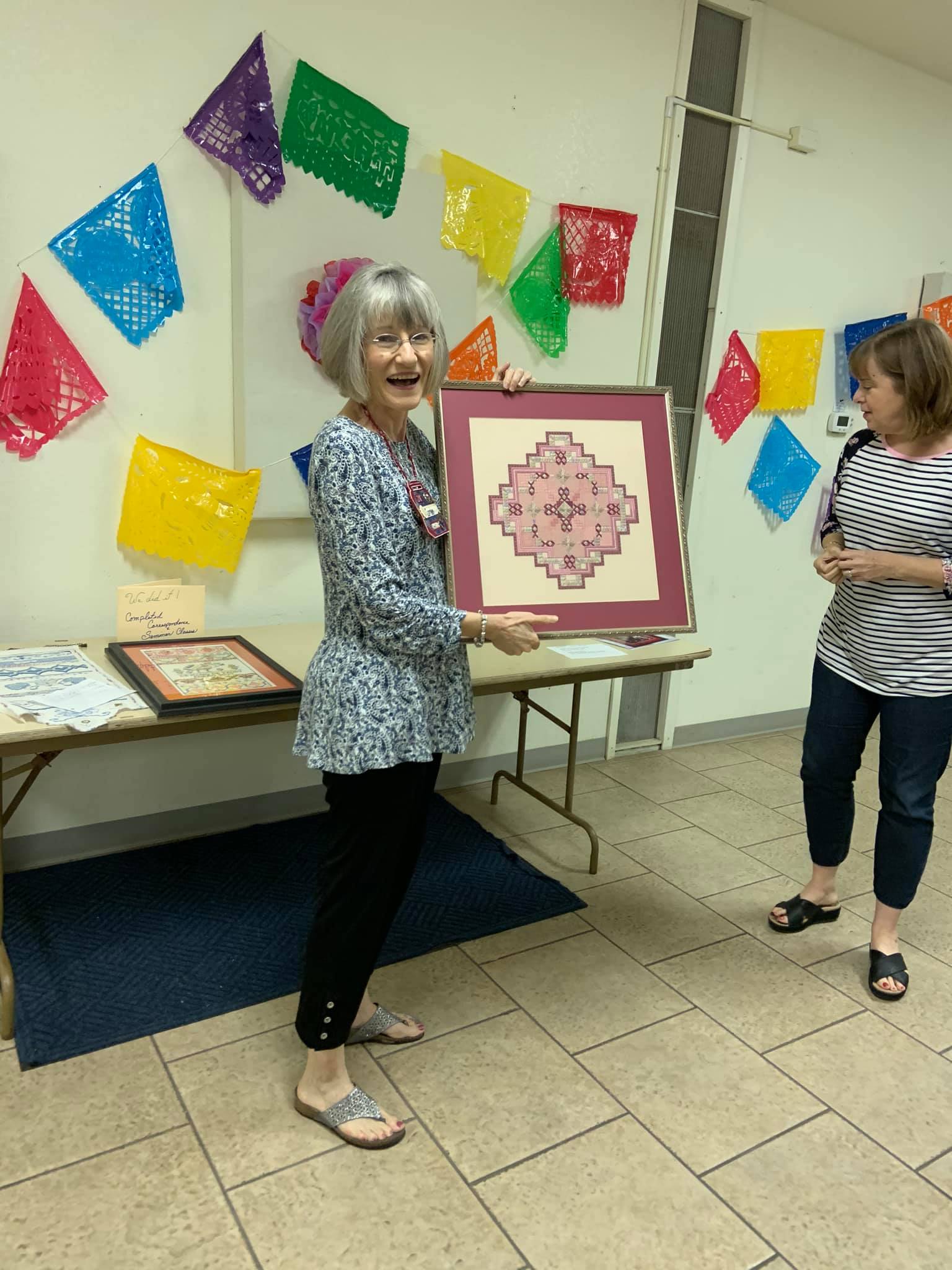 A person holds a framed pink needlepoint project while another person looks on in a room decorated with colorful bunting.