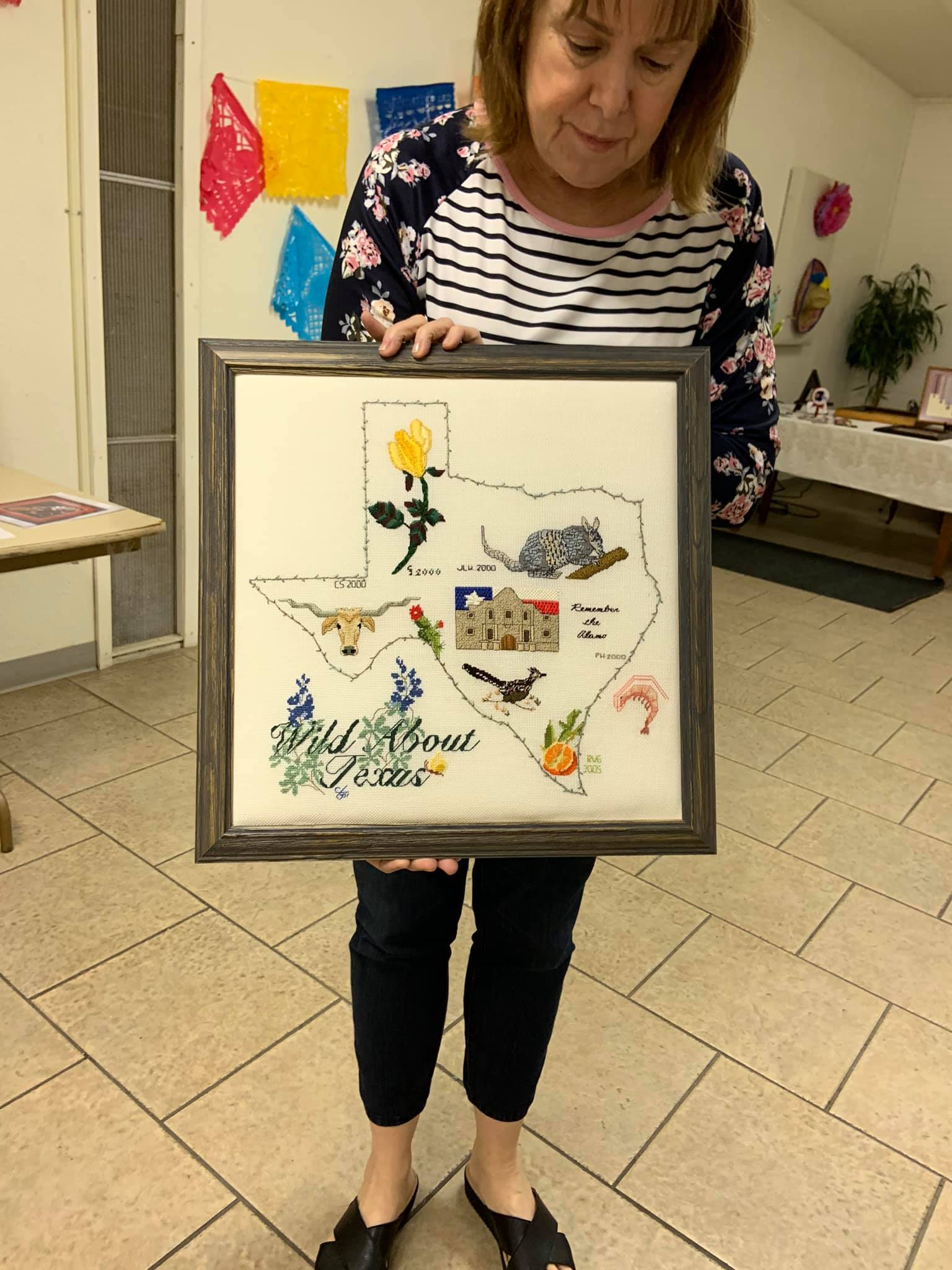 A person holds a framed cross-stitch map of Texas, decorated with iconic state symbols, in a bright, indoor setting.