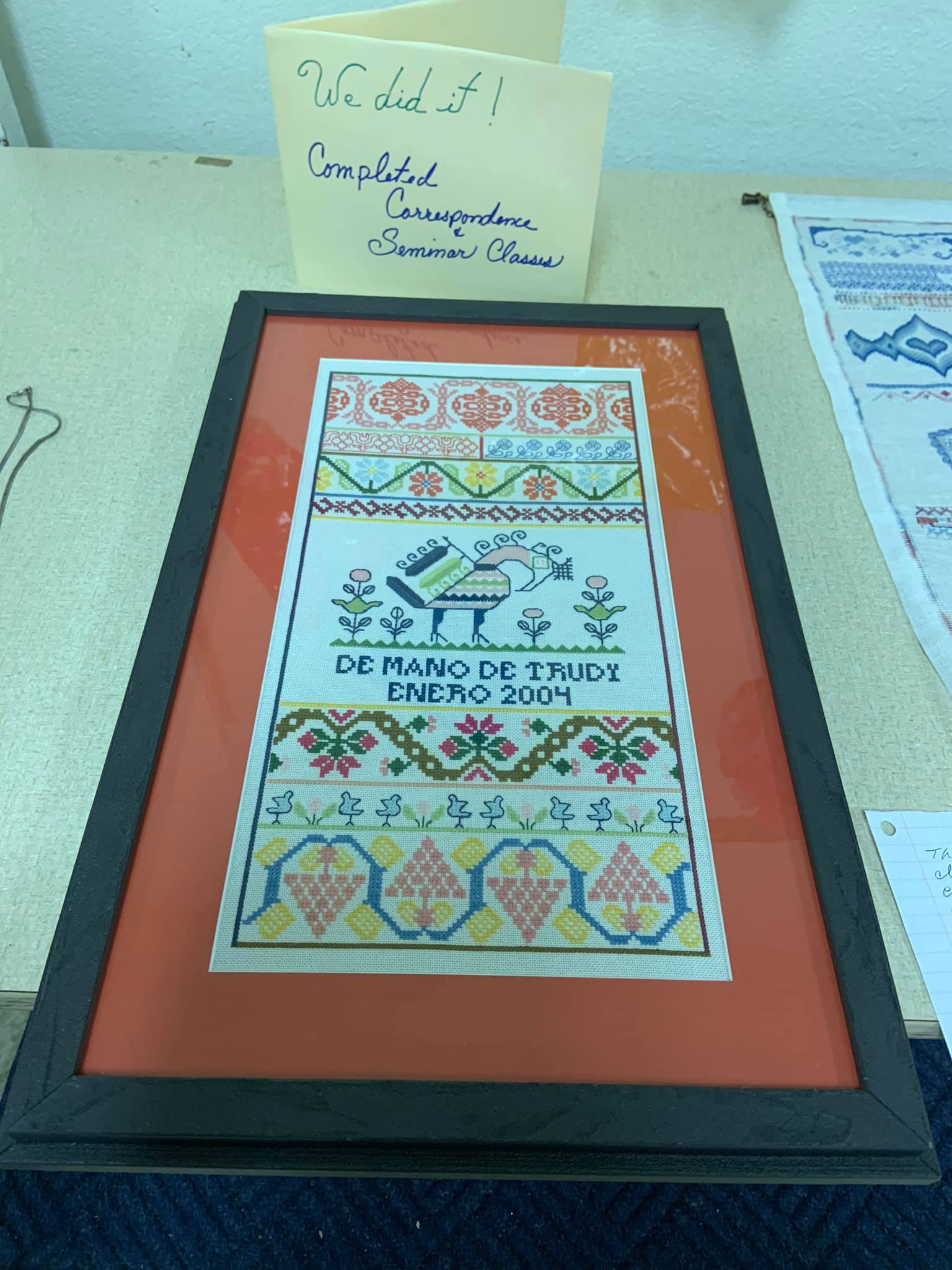 A framed, colorful cross-stitch sampler featuring a bird, floral patterns, and the text 