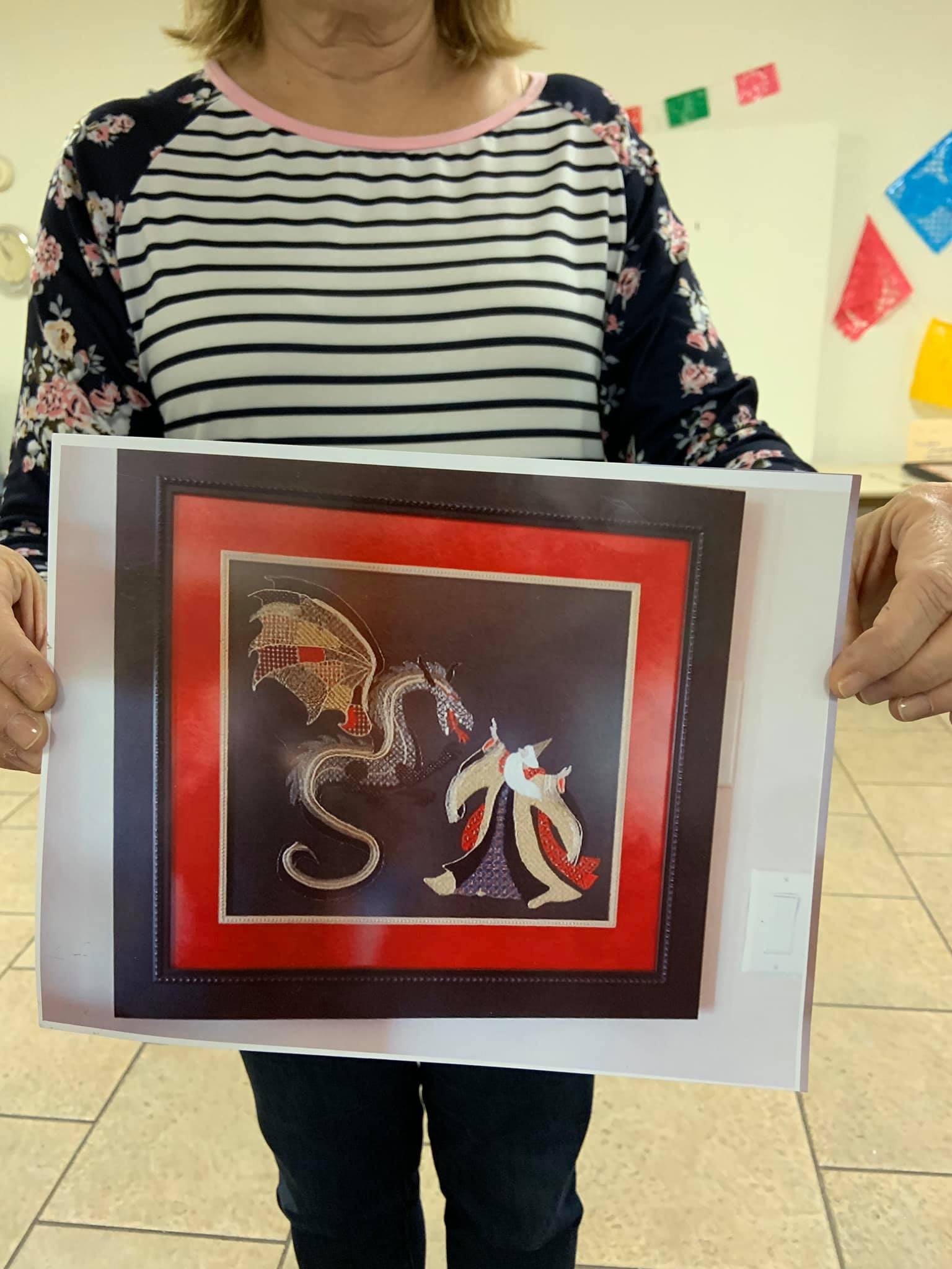 A person holds up a framed cross-stitch piece featuring a dragon and a figure in a red robe against a dark background.