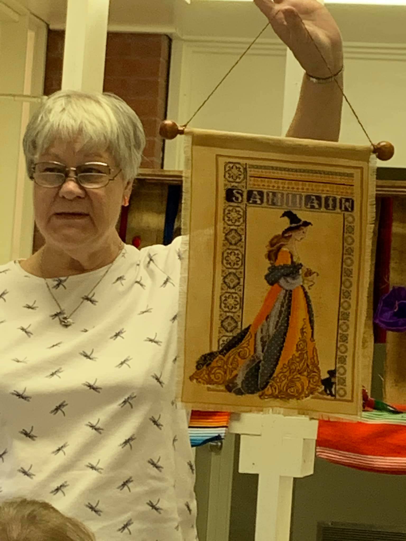 A person holds up a rectangular fabric wall hanging depicting a figure in a witch’s hat and an orange and black gown.