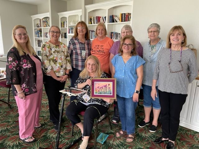 A group of nine people pose for a photo in a room with bookshelves, one holding a small framed piece of needlepoint.