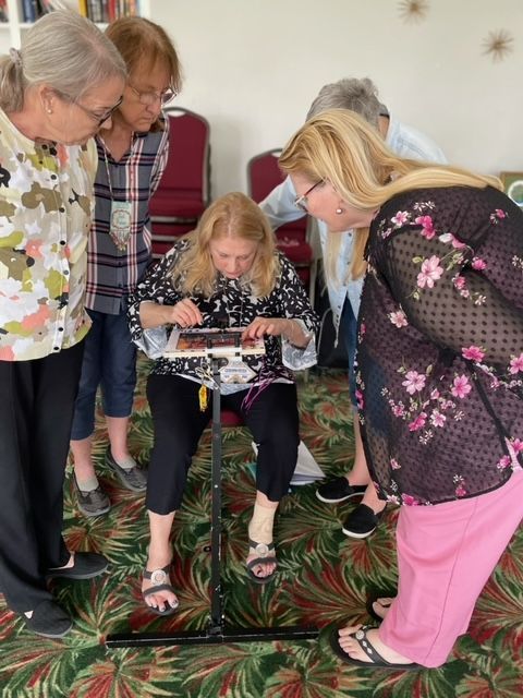 A woman sits and works on a needlepoint frame while four others stand around her, observing intently in a room.