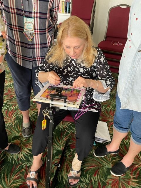 A person sits in a room working on embroidery on a stand, flanked by two others standing on either side.