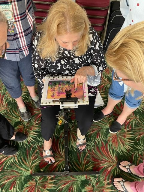 A person sits in a chair, pointing at a framed embroidery project on a stand while two others observe from above.