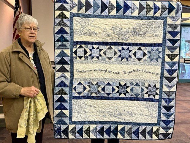 A person holds up a blue and white quilt with star patterns and inscribed text in an indoor setting with an American flag.
