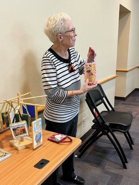 A person in a striped shirt stands by a table, holding up a small decorated craft item, with other crafts on display.