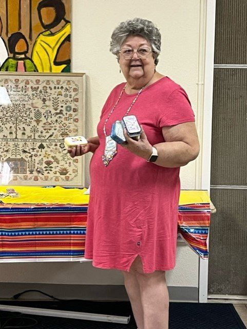 A smiling person in a pink dress stands by a table, holding a small white item in one hand and two tins in the other.