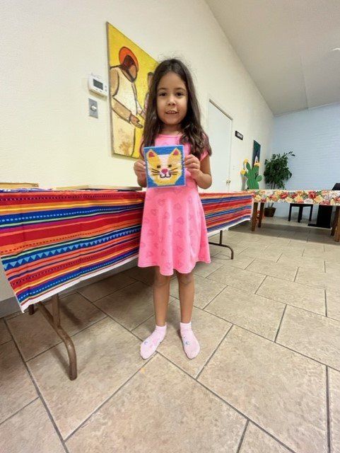 A child in a pink dress standing indoors, holding up a small square picture of a cartoon cat.