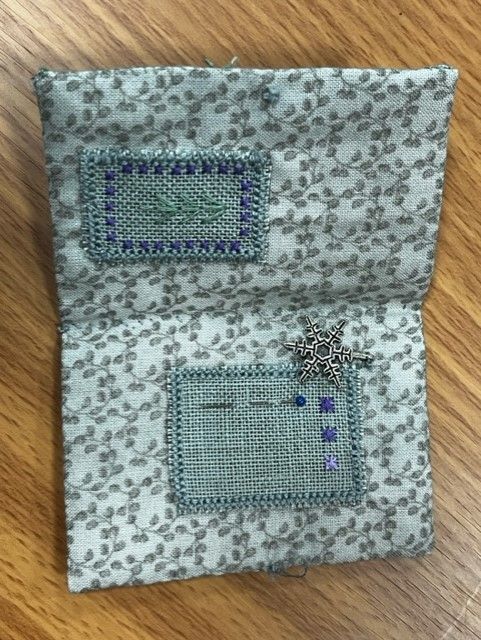 A folded fabric needle book with floral patterns, two stitched felt panels, a silver snowflake charm, and needles.
