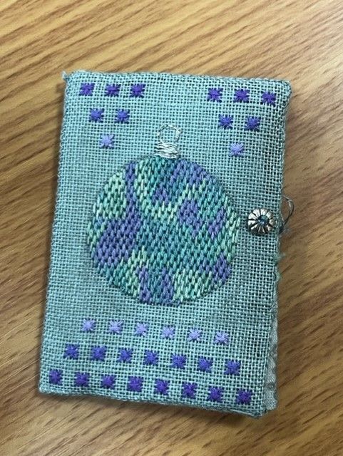 A small, embroidered fabric needle book with a blue-and-purple ornament design, featuring cross-stitched borders.